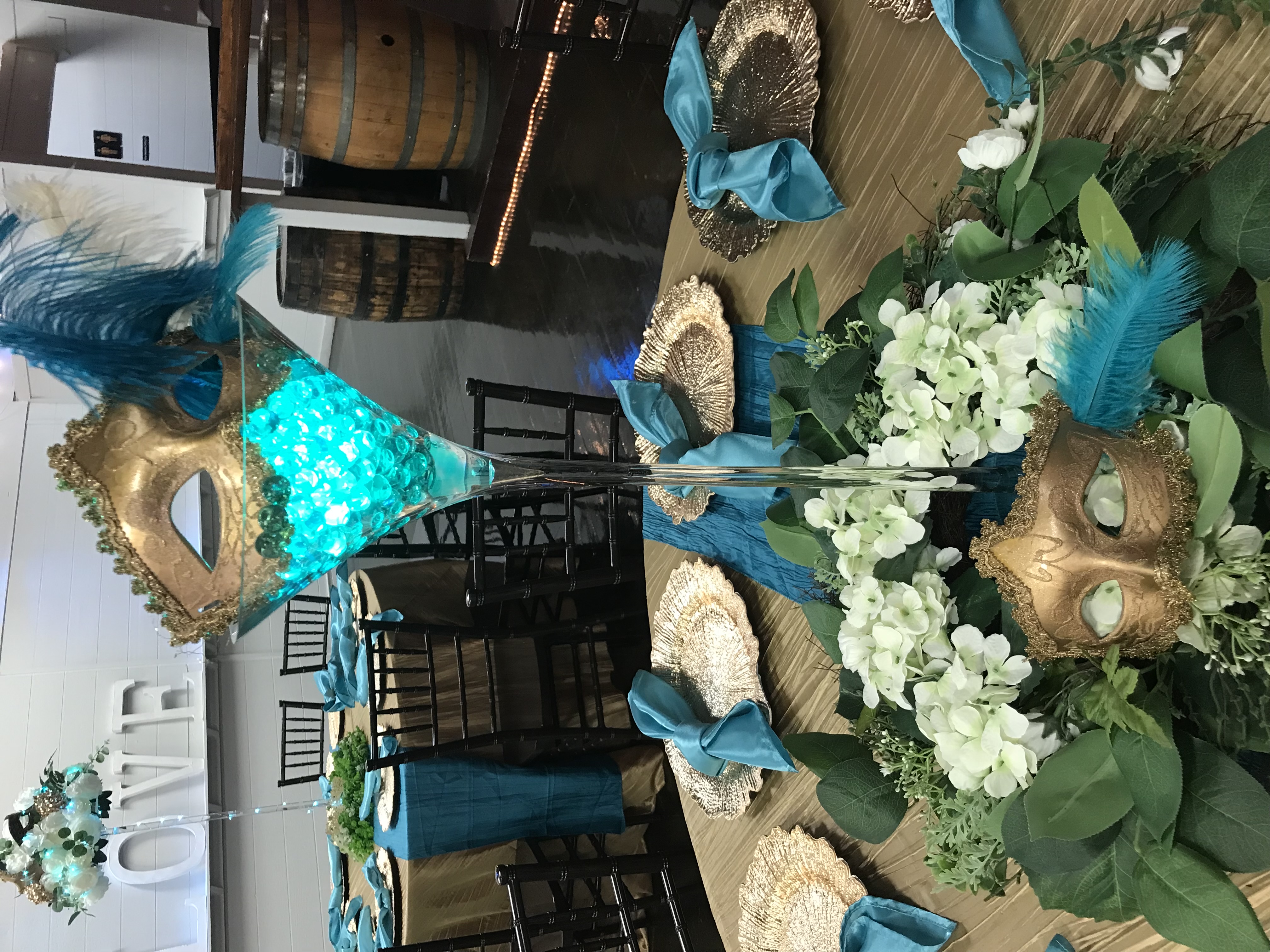 A masquerade ball centerpiece with flowers, greenery, feathers and a mask top this guest tables= with chargers and napkins.